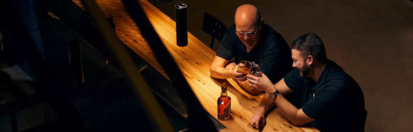 An overhead shot of two men, Fred Noe and Freddie Noe, smiling and sitting at a wooden bar, holding glasses of whiskey, with a bottle visible between them Whisky Hardin's Creek