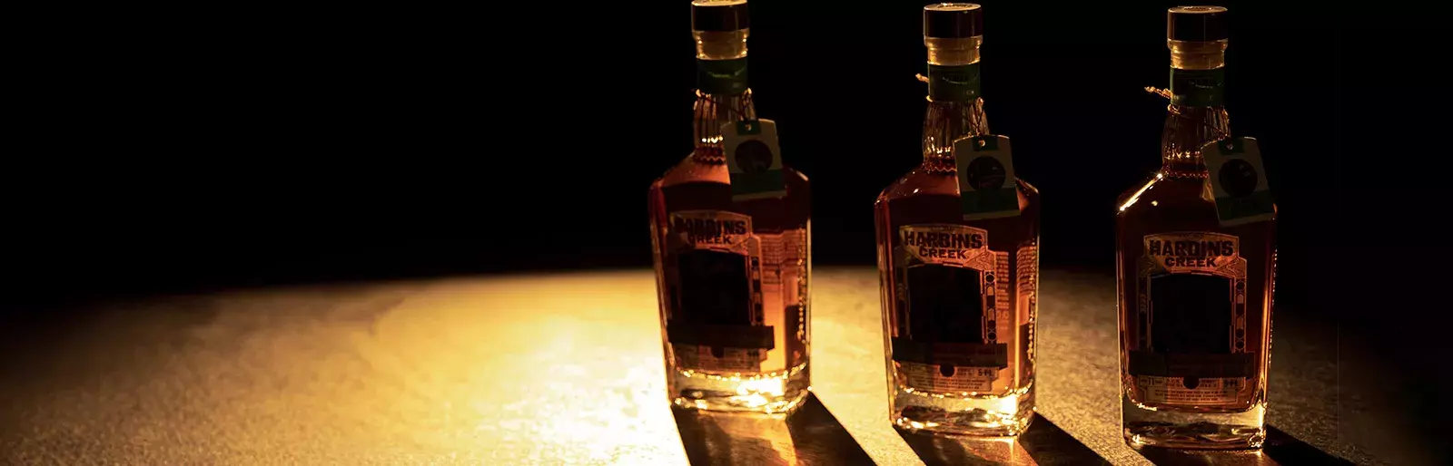Three whiskey bottles standing in a row on a dark, reflective surface, dramatically lit from the side to cast long, sharp shadows Whisky Hardin's Creek