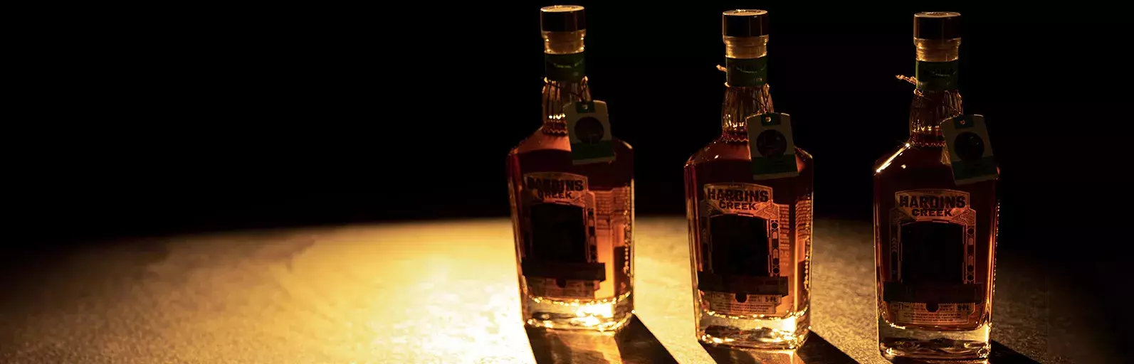 Three whiskey bottles standing in a row on a dark, reflective surface, dramatically lit from the side to cast long, sharp shadows Whisky Hardin's Creek