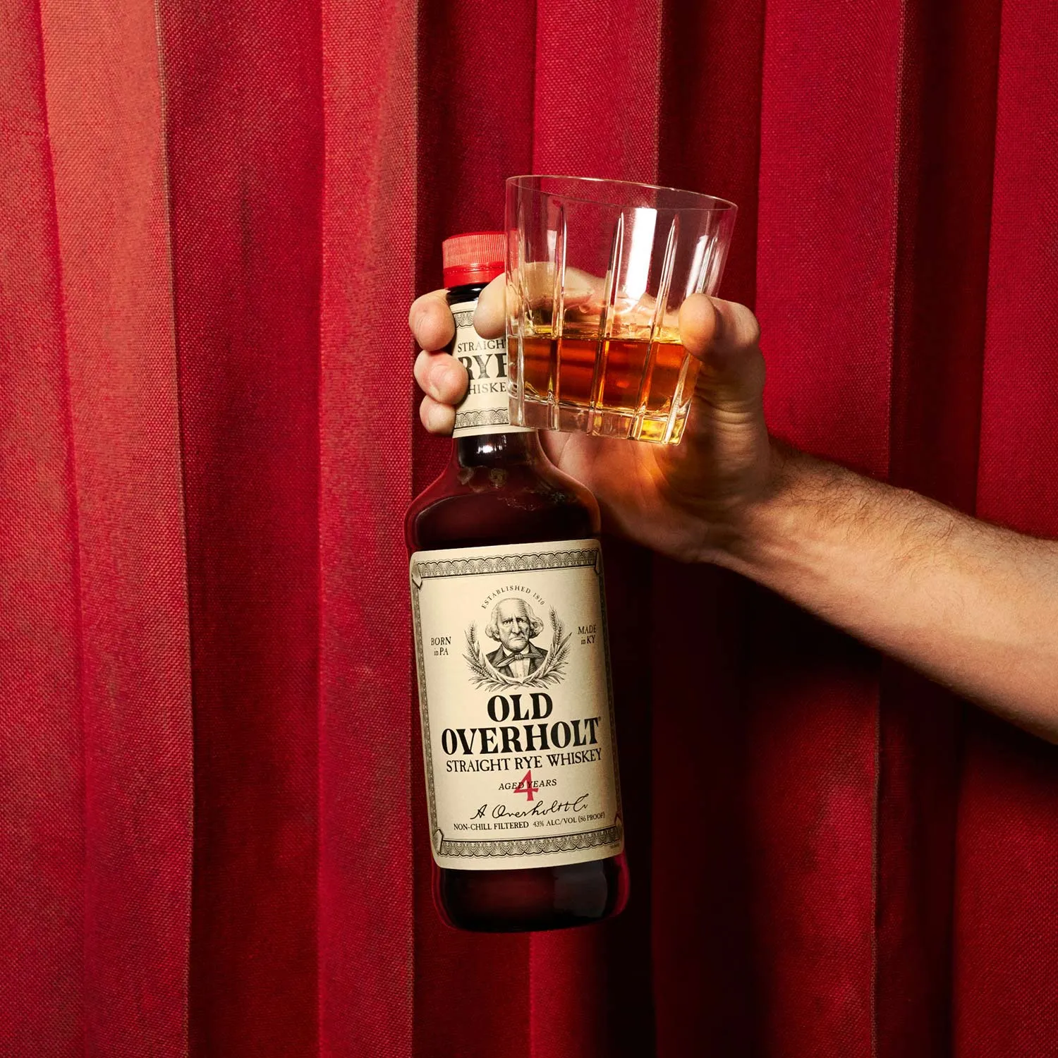 Old one cold one cocktal with hand holding a glass of whiskey next to a bottle of American Whisky Old Overholt.