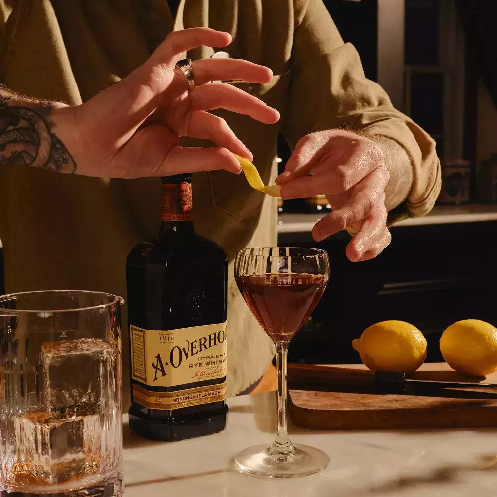 A person's hands garnish a cocktail with a lemon twist, next to a bottle of American Rye Whiksy Old Overholt