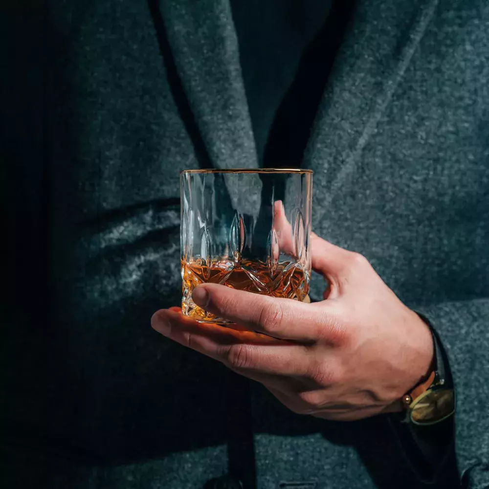 A close-up of a man's hand, wearing a wristwatch, holding a gold-rimmed cut-crystal glass of whiskey against his dark gray coat Whisky Hardin's Creek