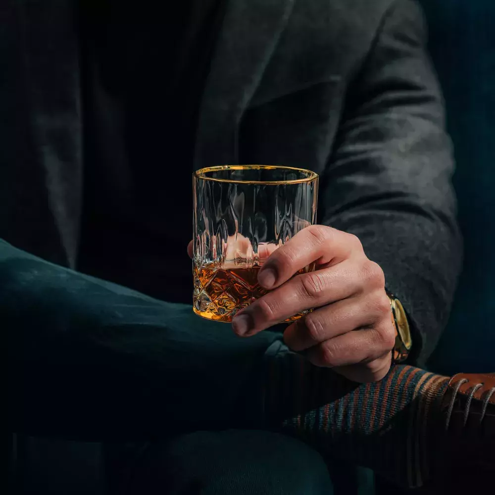 A man in a dark coat and trousers holds a faceted glass of whiskey, revealing a gold wristwatch and striped socks near his shoe Whisky Hardin's Creek