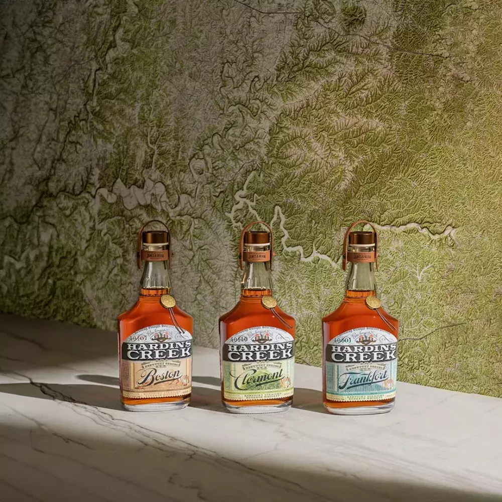 Three bell-shaped bottles of the Kentucky Series (Boston, Clermont, Frankfort) standing in a row against a textured green and white topographical map background Whisky Hardin's Creek