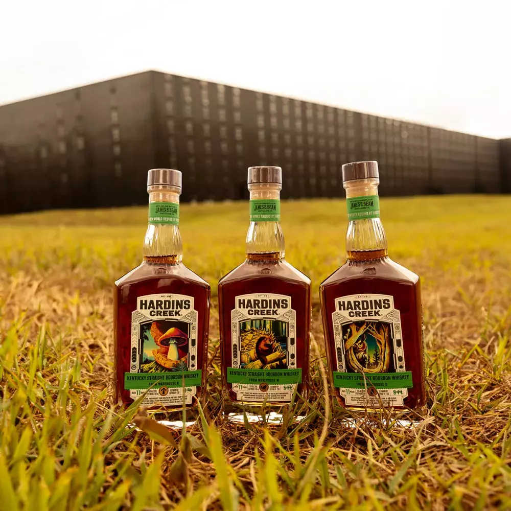 Three square-shouldered bourbon bottles (The Mushroom, The Beaver, and The Owl) standing in tall grass in front of a massive, dark bourbon rickhouse Whisky Hardin's Creek.