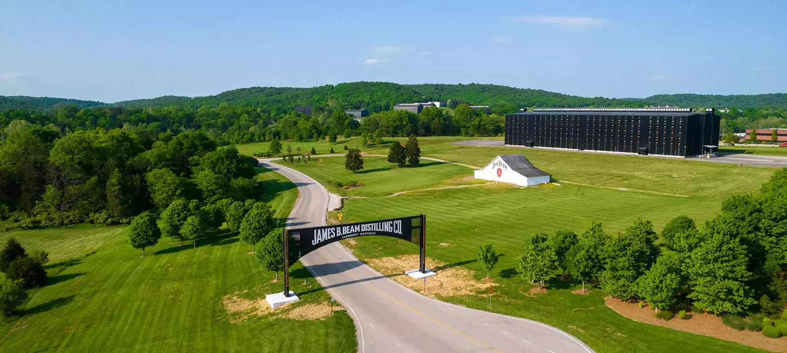 An aerial view of the James B. Beam Distilling Co. campus, showing a road, green fields, a white house, and a large, black rickhouse building Whisky Hardin's Creek