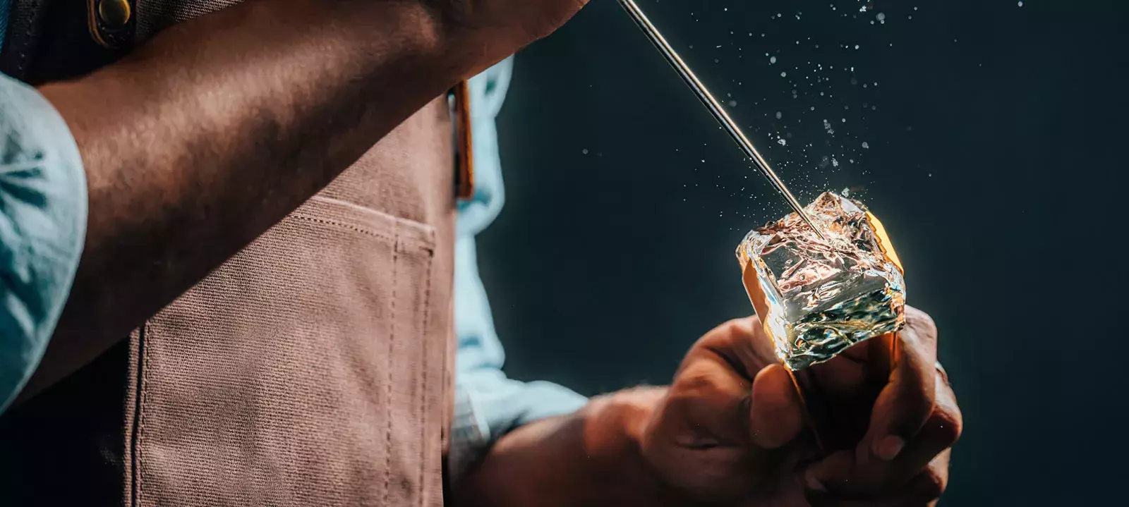 A close-up of a person wearing a brown apron using a metal pick to chip away at a large, clear block of ice Whisky Hardin's Creek