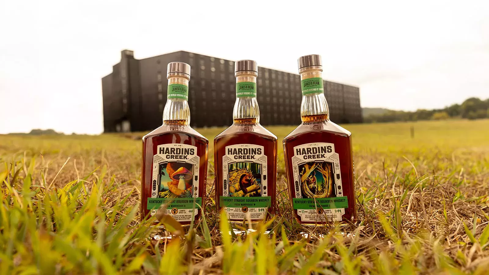 Three square-shouldered bourbon bottles (The Mushroom, The Beaver, and The Owl) standing in tall grass in front of a massive, dark bourbon rickhouse Whisky Hardin's Creek.  Development CTA: L