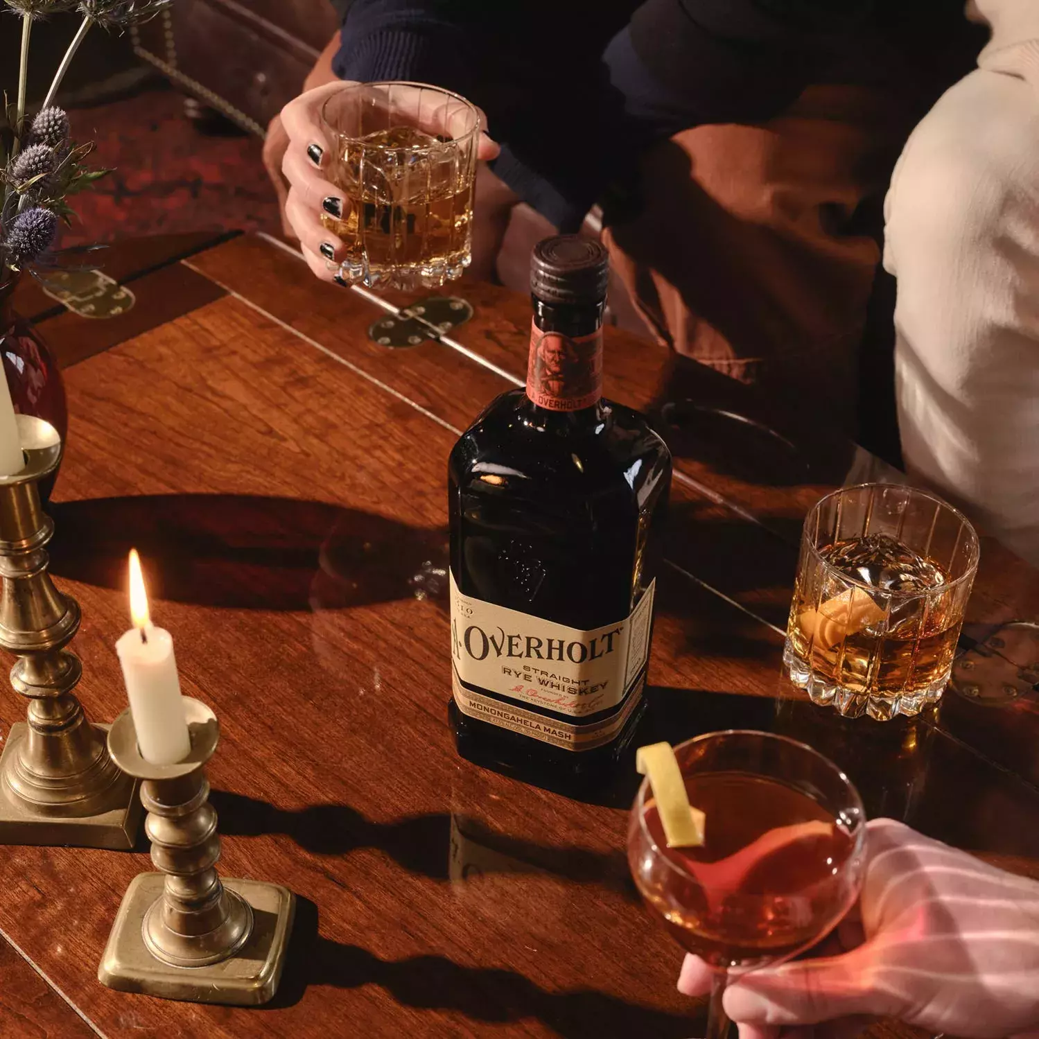 Hands holding glasses with cocktails and whiskey around a bottle of Monongahela Mash Straight Rye American Whisky Old Overholt.