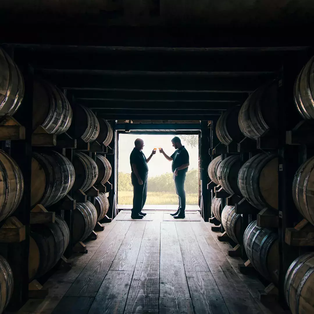 Two figures silhouetted in a bright doorway at the end of a dark bourbon rickhouse aisle lined with barrels, toasting glasses of whiskey Whisky Hardin's Creek