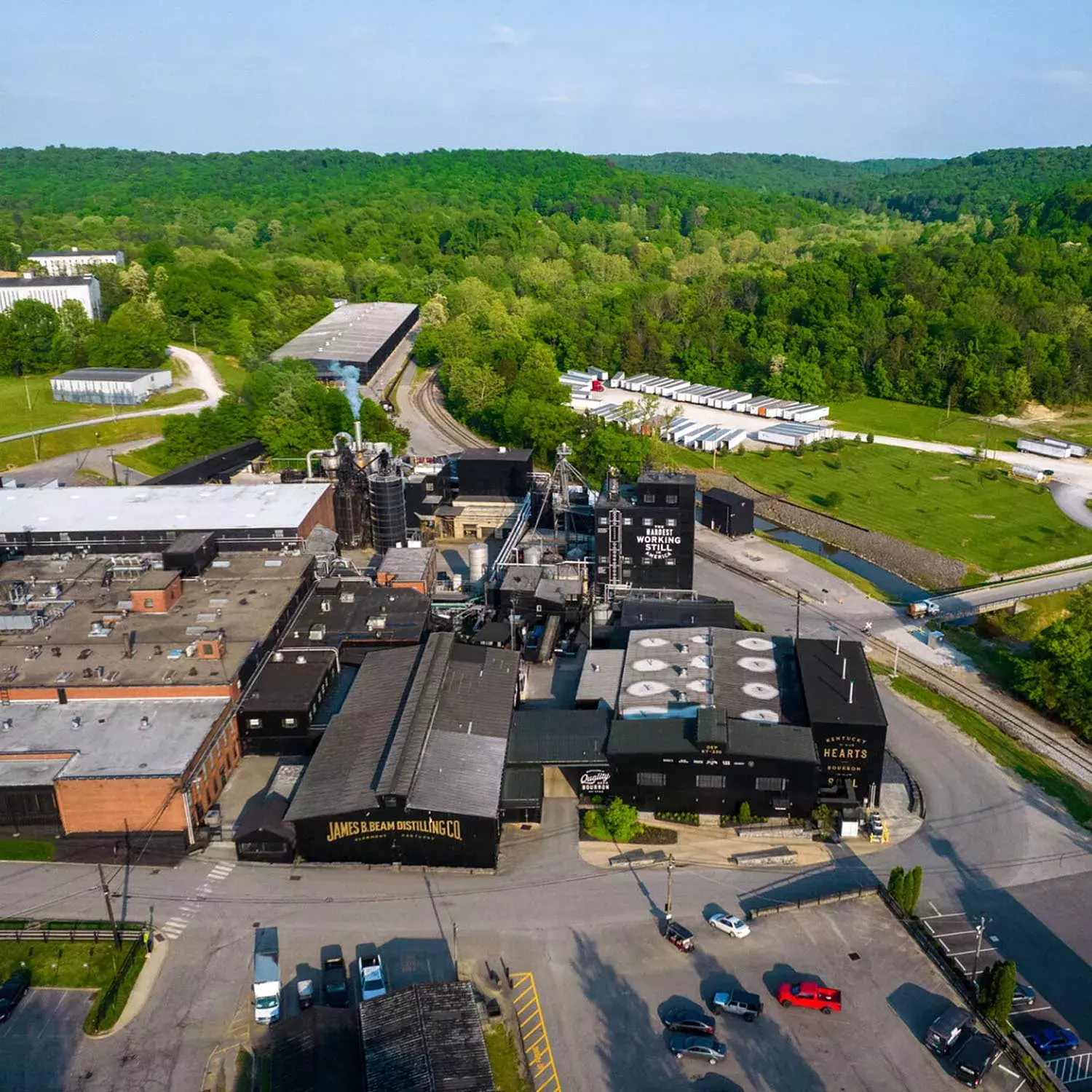 An aerial view of the James B. Beam Distilling Co. facility at Clermont, Kentucky, showing a complex of industrial buildings, black warehouses, a parking lot, and surrounding green hills and forest Whisky Hardin's Creek