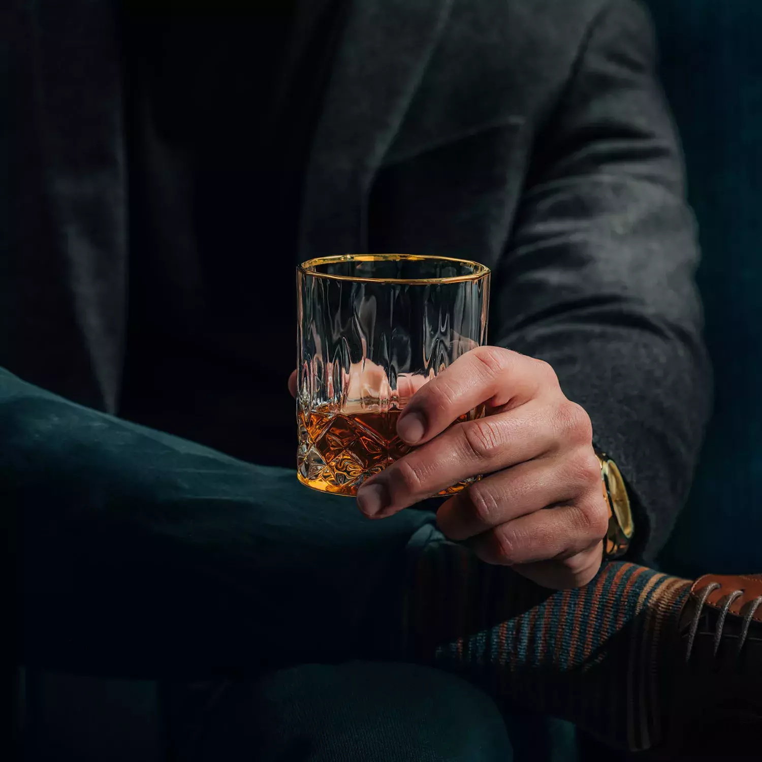 A man in a dark coat and trousers holds a faceted glass of whiskey, revealing a gold wristwatch and striped socks near his shoe Whisky Hardin's Creek