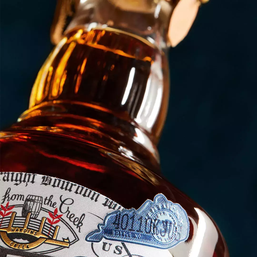 A macro shot of a whiskey bottle label, focusing on the dark amber liquid, the text, and a blue and white Kentucky state tax stamp Whisky Hardin's Creek