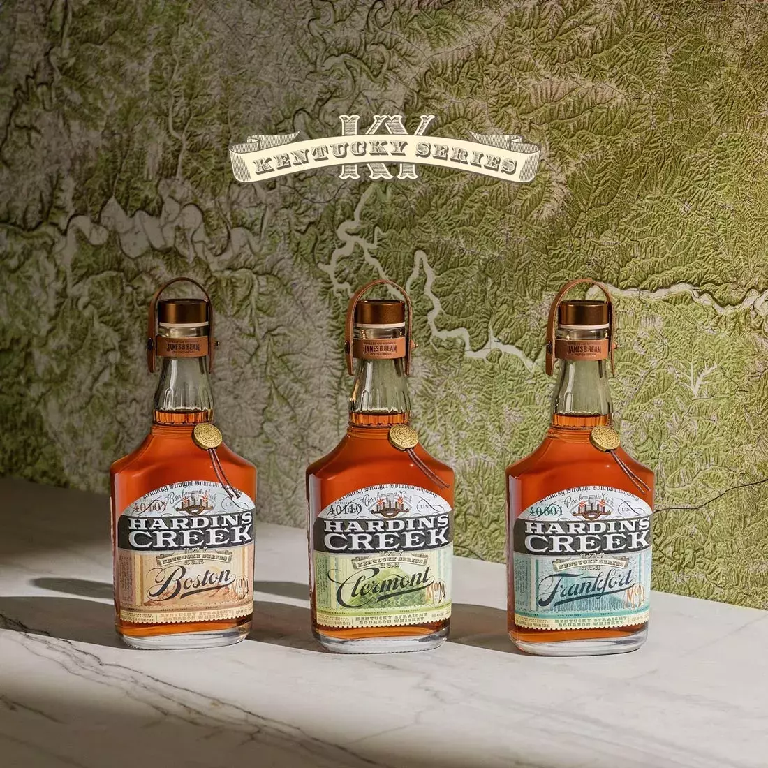 Three bell-shaped bottles of the Kentucky Series (Boston, Clermont, and Frankfort) standing in a row against a textured green and white topographical map background with a banner reading "Kentucky Series" Whisky Hardin's Creek