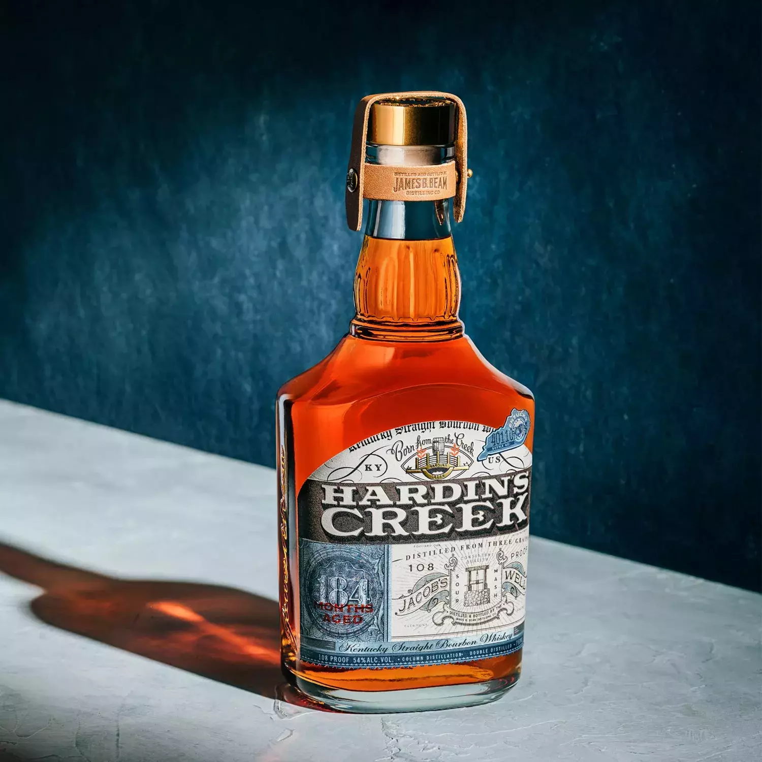 A bell-shaped bottle of the Jacob's Well expression, aged 184 months, standing on a light surface against a dark blue background with harsh lighting Whisky Hardin's Creek