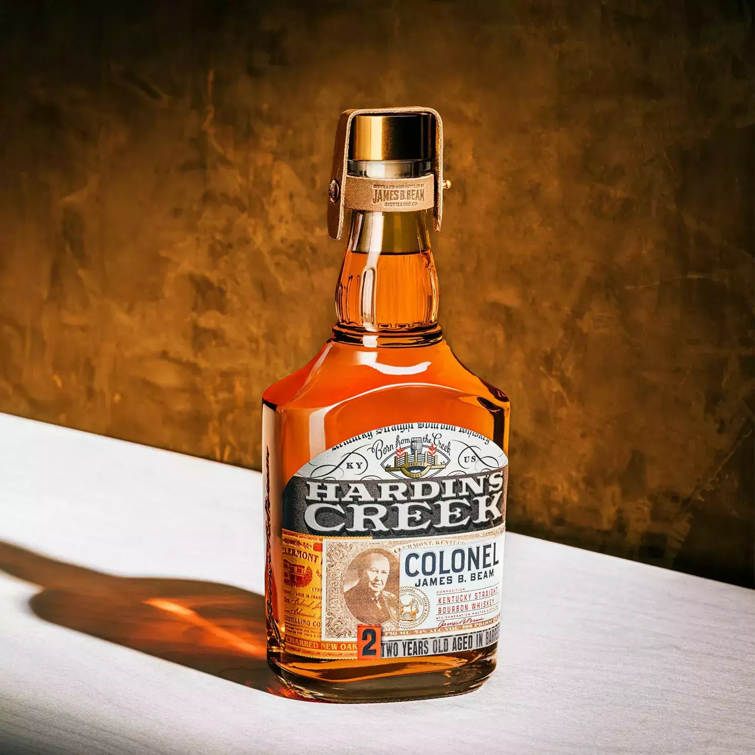 A bell-shaped bottle of the Colonel expression, aged two years, standing on a light surface against a gold background with harsh lighting Whisky Hardin's Creek