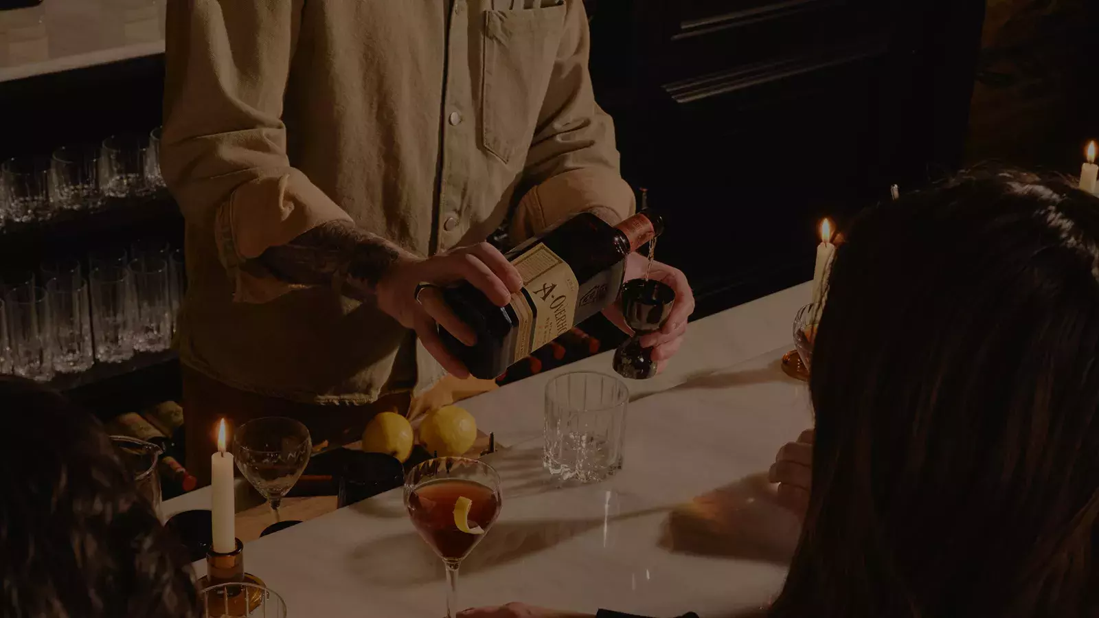 Hands pouring whiskey from a bottle into a jigger, with a glass and lemons on a bar, featuring American Whisky Old Overholt.