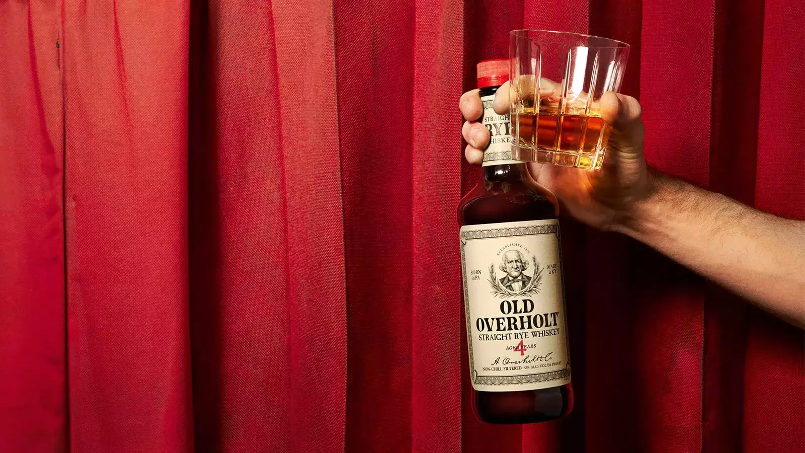 Old one cold one cocktal with hand holding a glass of whiskey next to a bottle of American Whisky Old Overholt.