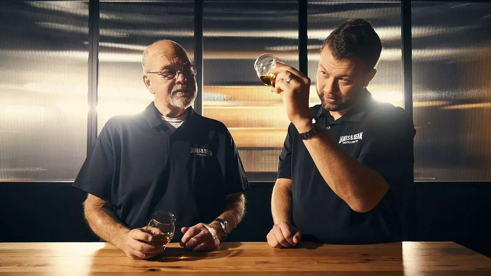 Fred Noe and Freddie Noe in black shirts sitting at a wooden table, examining glasses of amber whiskey in bright, dramatic light Whisky Hardin's Creek