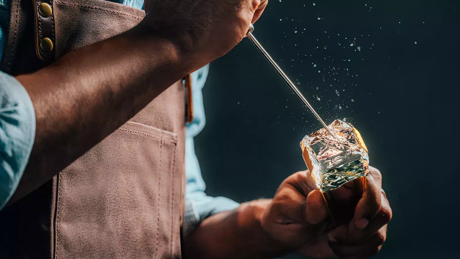 A close-up of a person wearing a brown apron using a metal pick to chip away at a large, clear block of ice Whisky Hardin's Creek