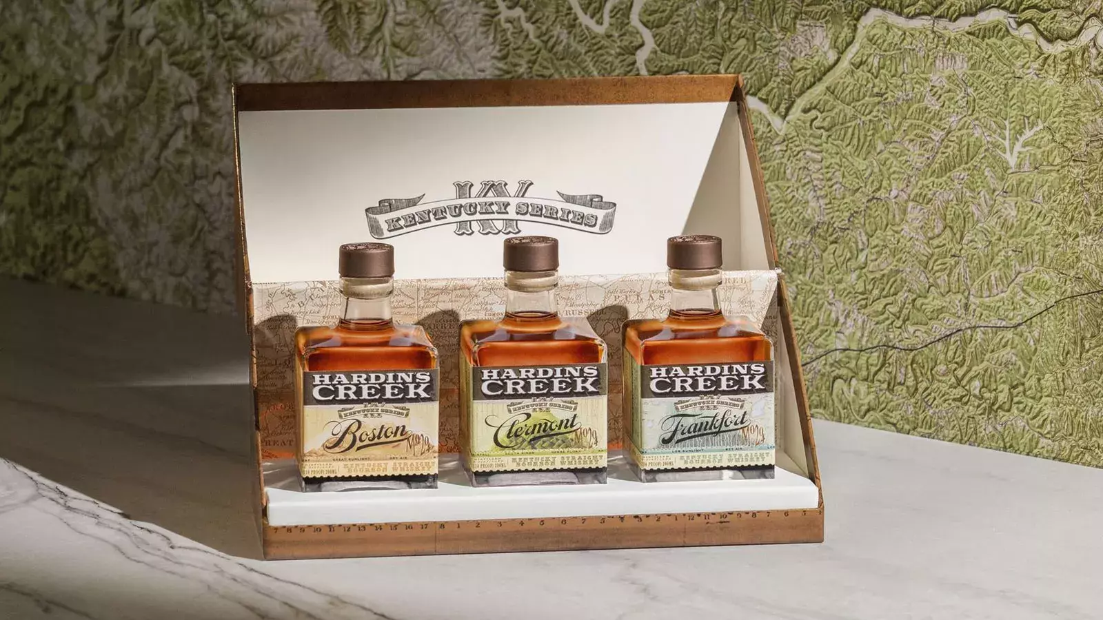 A presentation box displaying three smaller, bell-shaped bottles labeled Boston, Clermont, and Frankfort from the "Kentucky Series" Whisky Hardin's Creek.