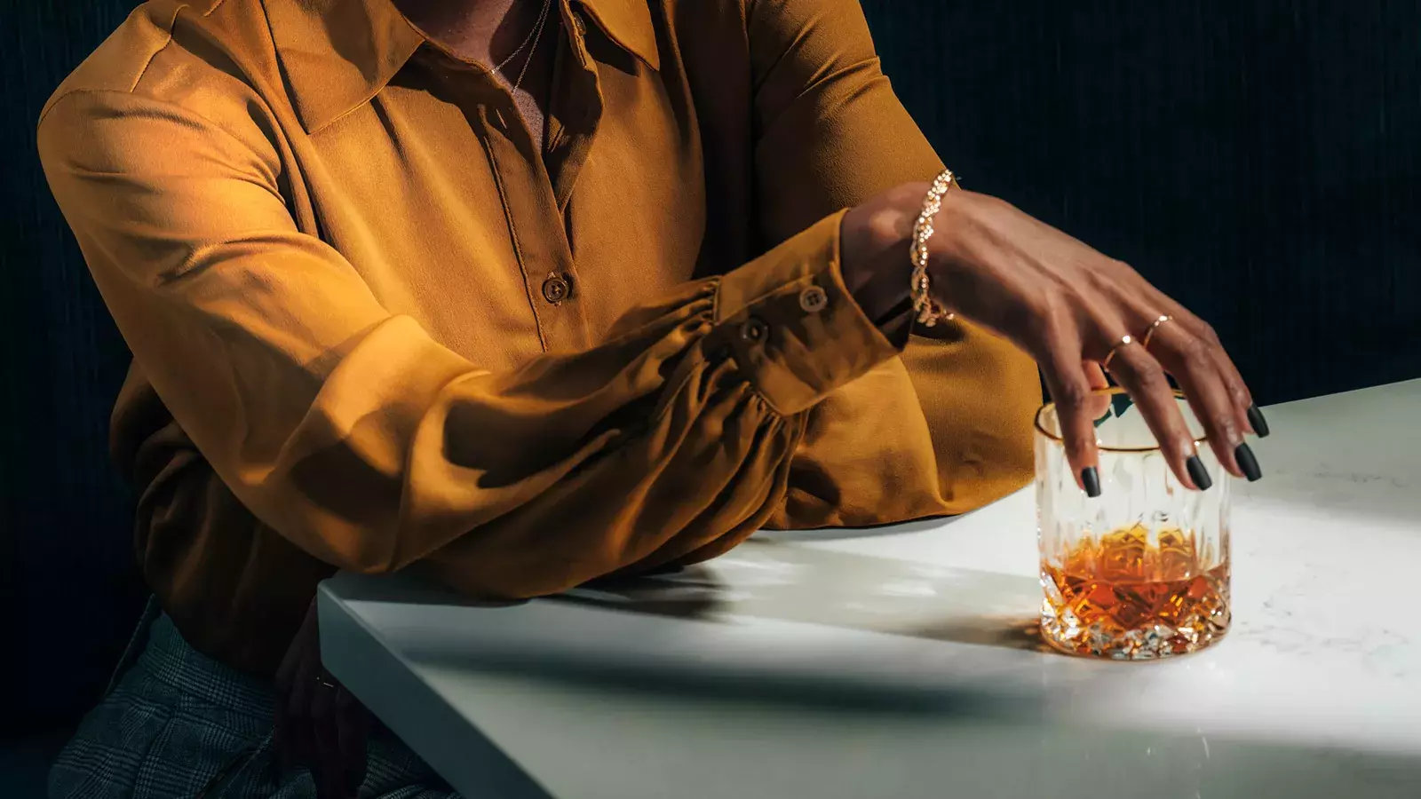 A close-up of a woman's hands holding a short whiskey glass with an amber drink and a large spherical ice ball, showing her matte black fingernails Whisky Hardin's Creek.