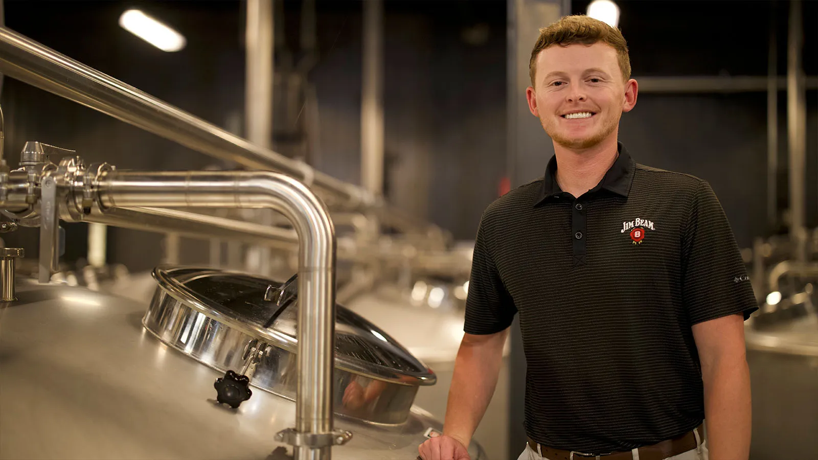 Smiling distiller Alex Mayes from James B. Beam Distilling Co. standing next to stainless steel fermentation vats. Bourbon making.