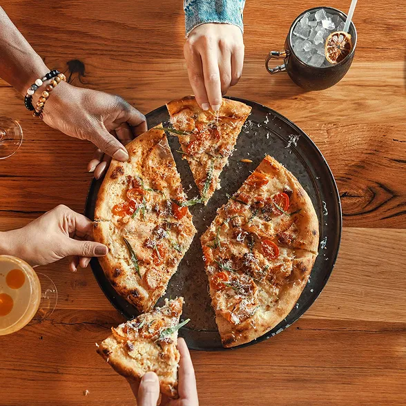 Hands reaching for slices of a wood-fired pizza at The Kitchen Table restaurant.  Development