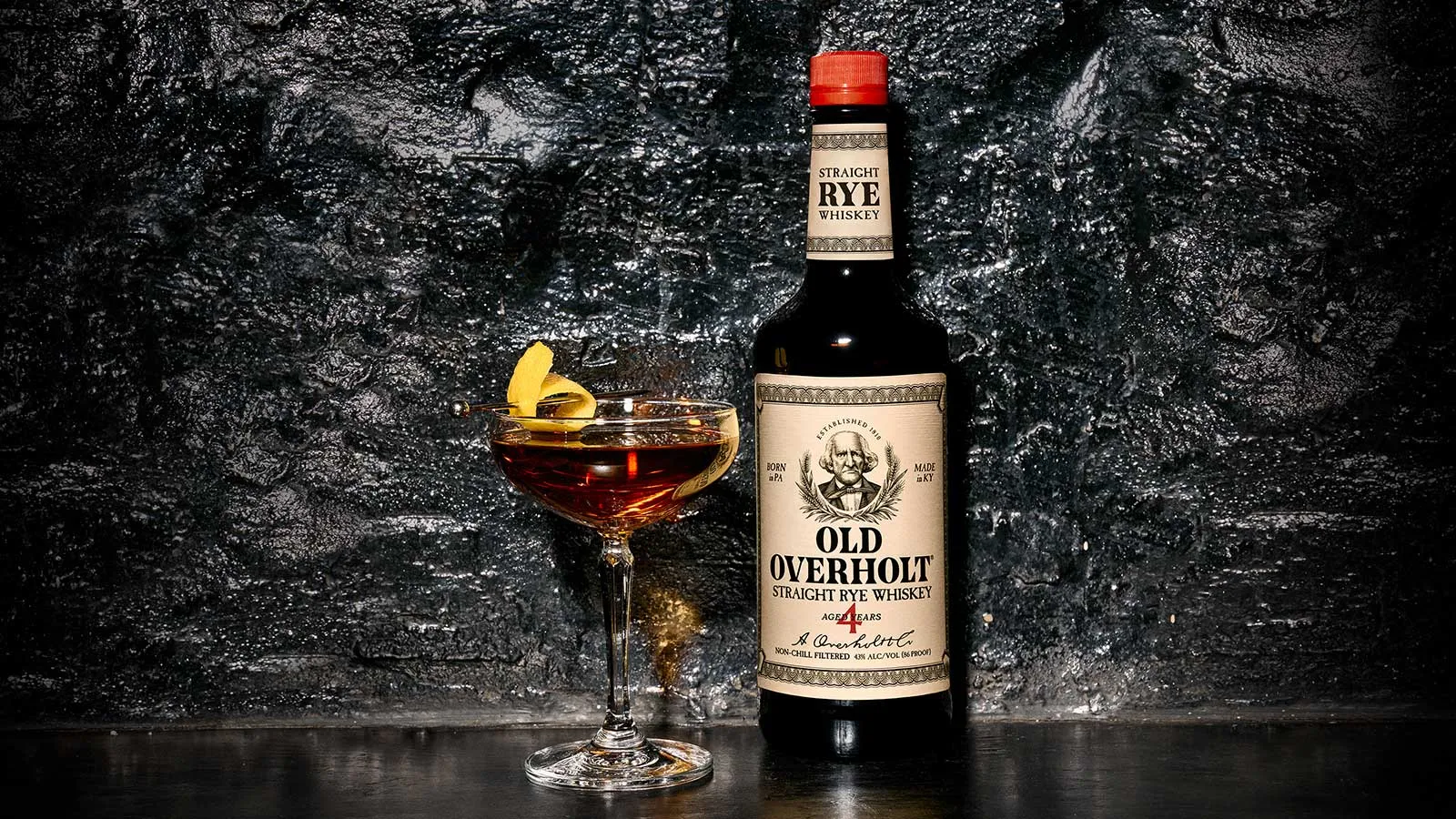 A cocktail with a lemon peel next to a bottle of Old Overholt 4 Year Straight Rye American Whisky Old Overholt.