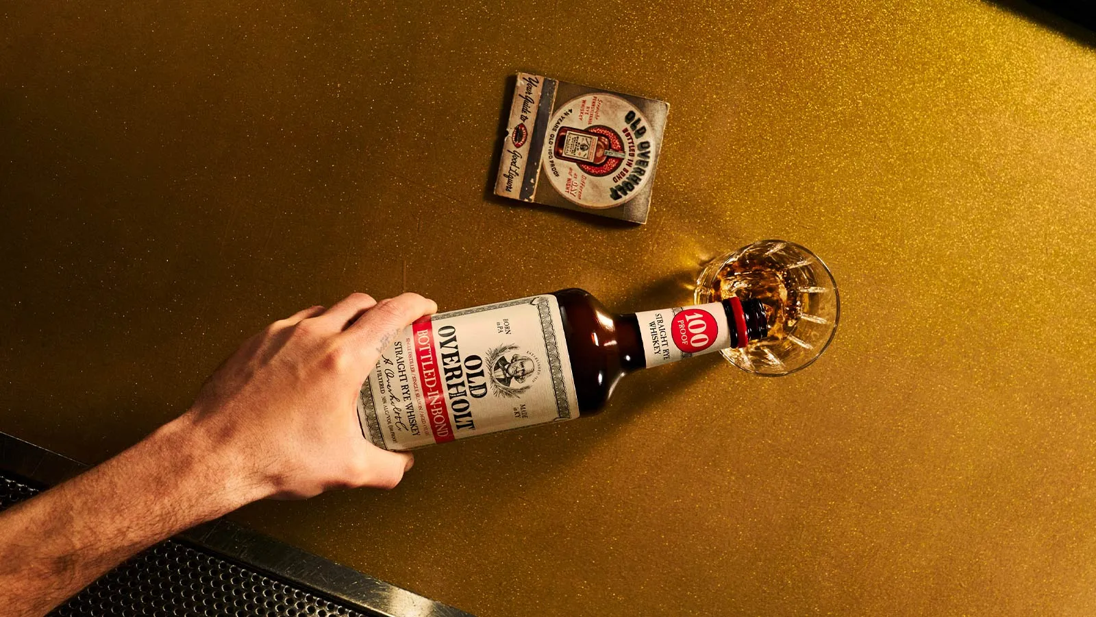 Hand pouring Old Overholt Bottled-in-Bond rye into a glass, with a matchbook on a gold surface, American Whisky Old Overholt.