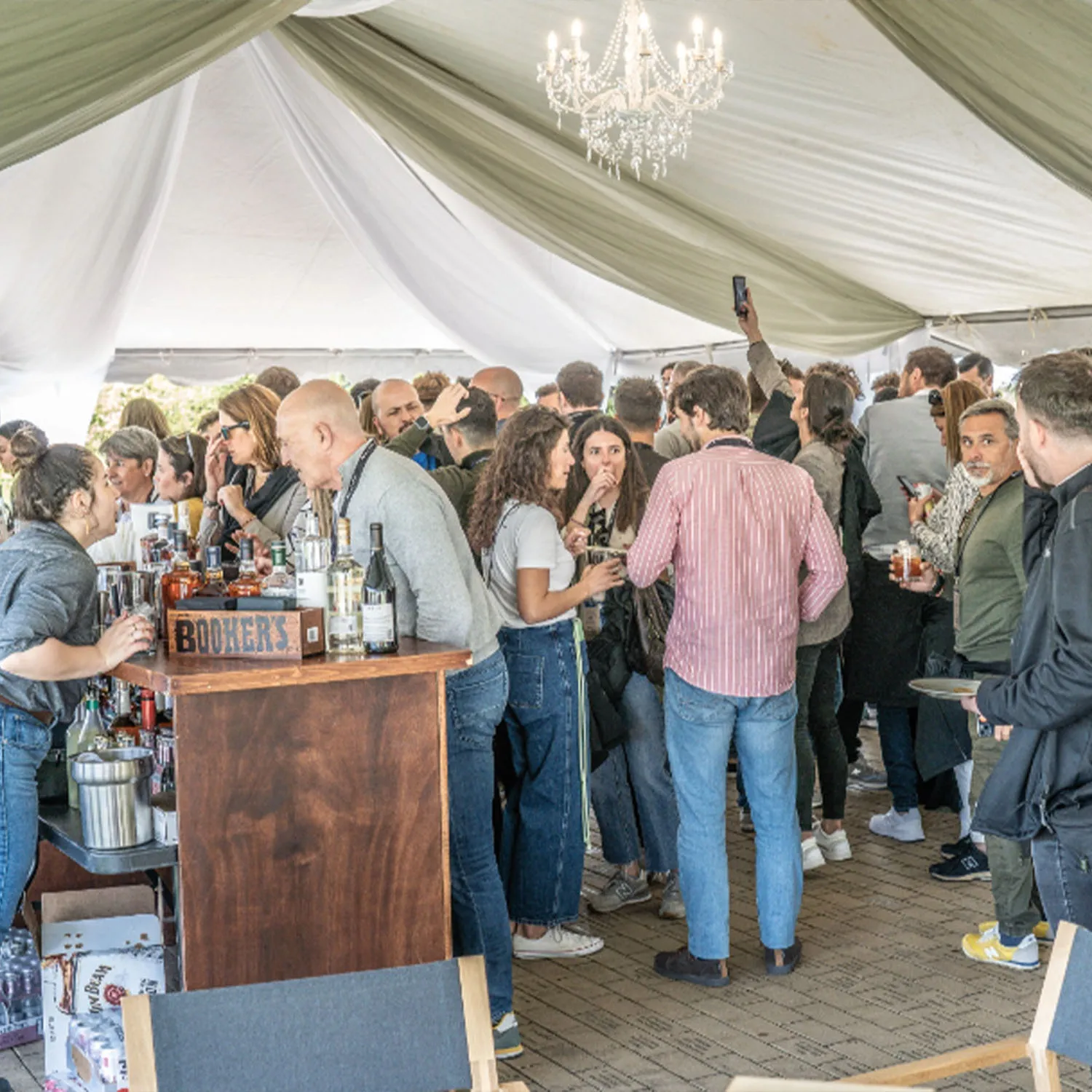 Crowd socializing at a bar under a tent with a chandelier at a James B Beam Distilling Co. event.