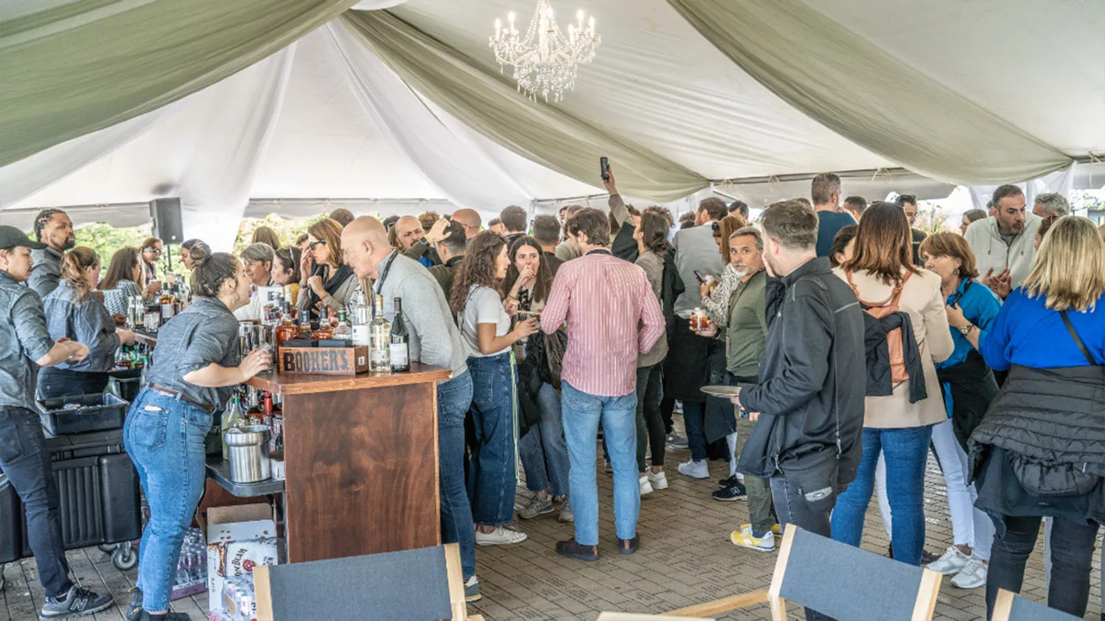 Crowd socializing at a bar under a tent with a chandelier at a James B Beam Distilling Co. event.