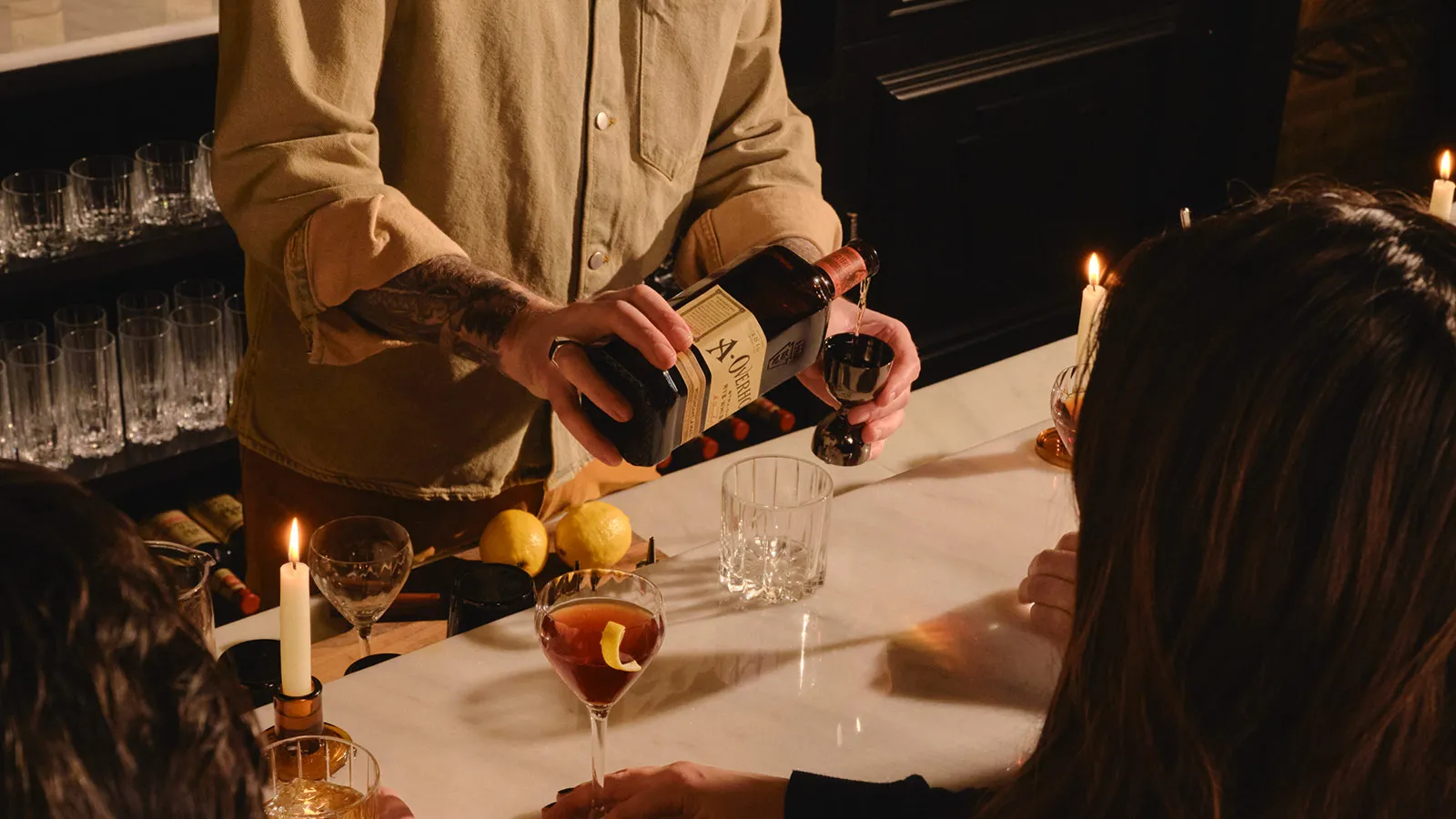 Hands pouring whiskey from a bottle into a jigger, with a glass and lemons on a bar, featuring Monongahela Mash Straight Rye American Whisky Old Overholt.