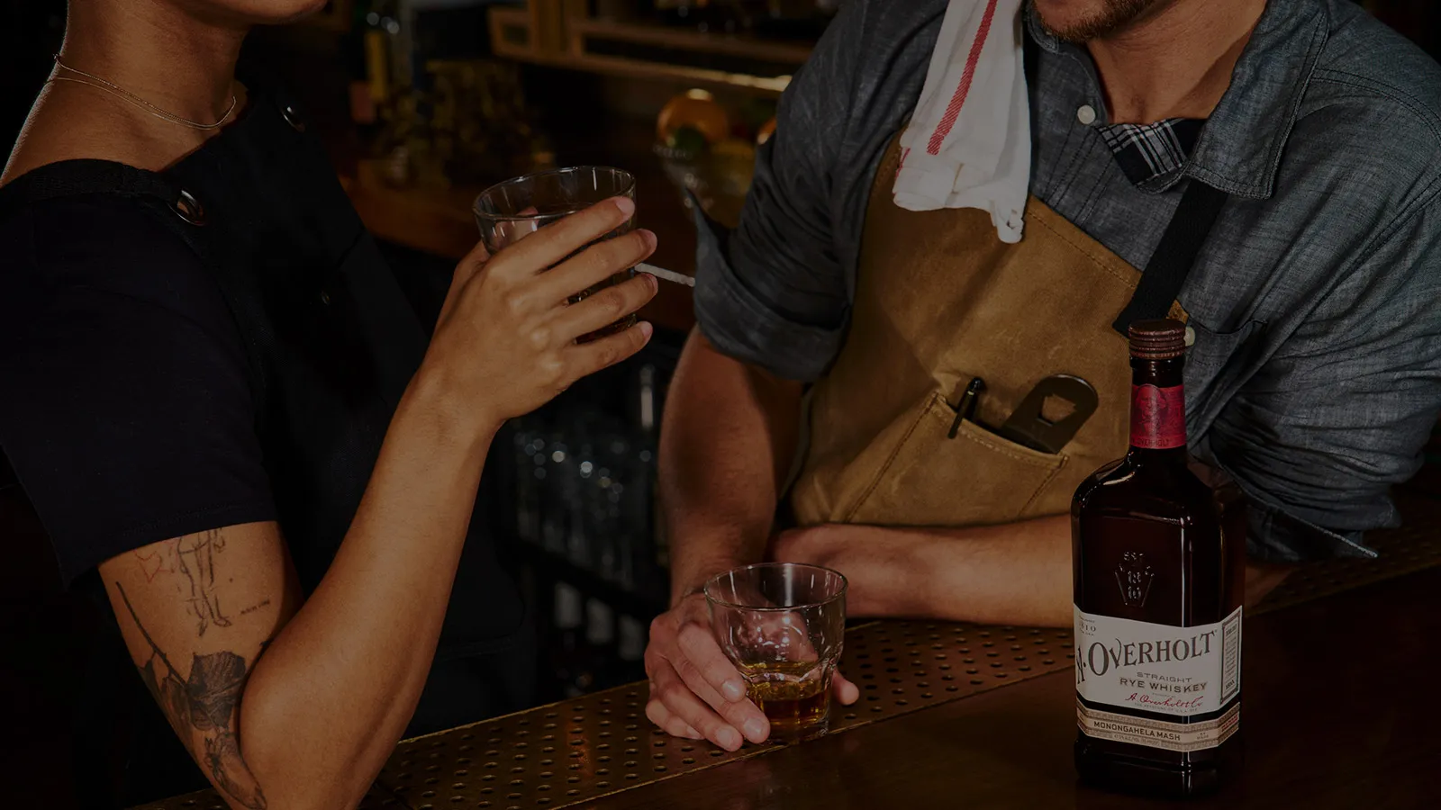 Two people at a bar with glasses of whiskey and a bottle of Monongahela Mash Straight Rye American Whisky Old Overholt.